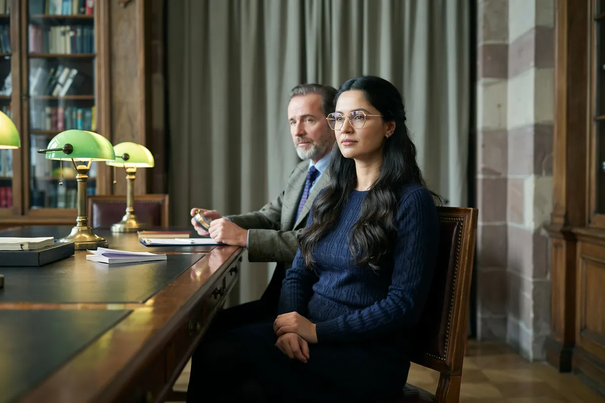 Frau mit langem dunklem Haar und runder Brille sitzt konzentriert in einer historischen Bibliothek, ihr Coach sitzt neben ihr