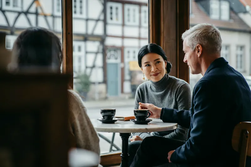 Frau mit tiefem Haarknoten lächelt am Marmortisch eines Cafés ihrem Coach zu, dazwischen Espresso und ein Notizbuch