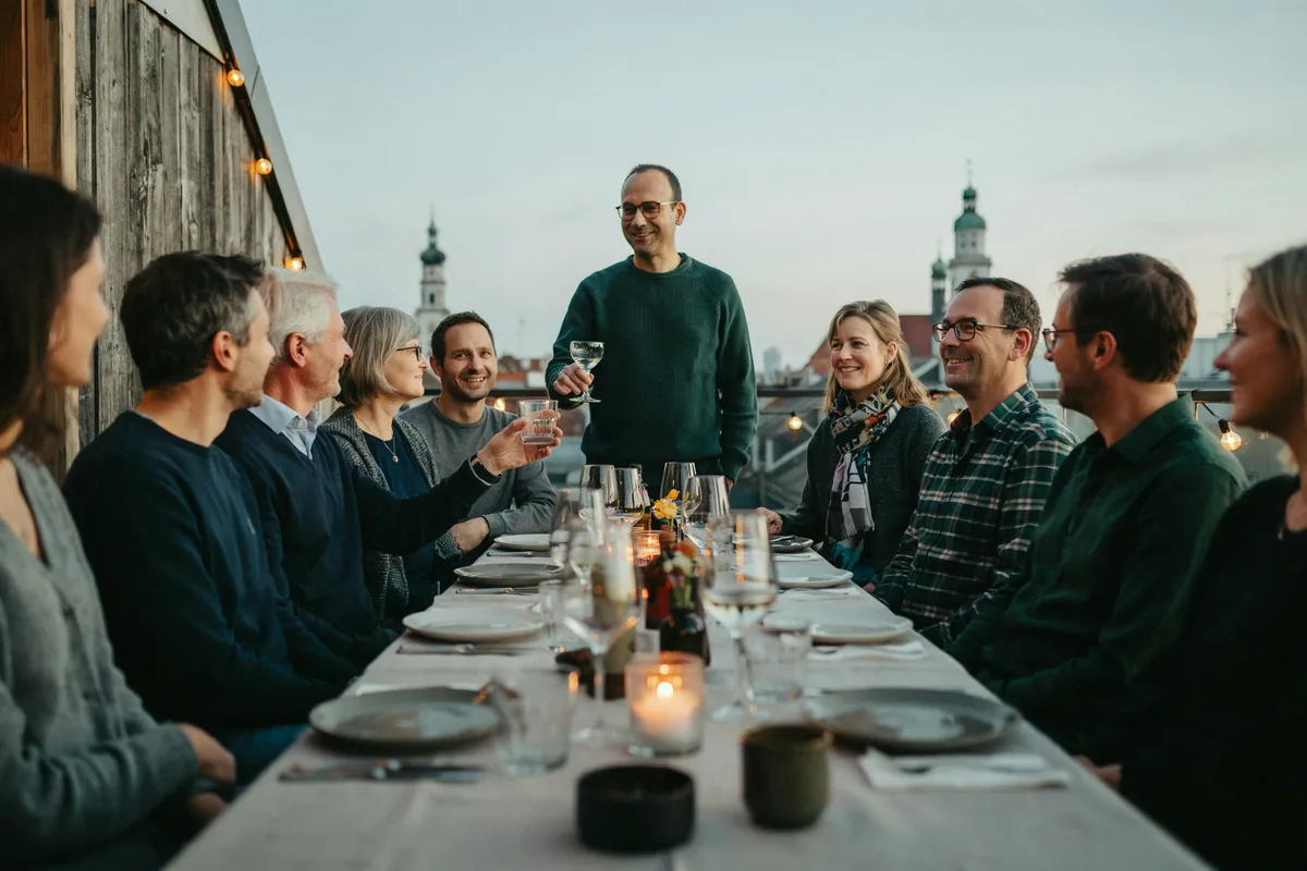 Social Media Relationship Manager: Community pflegen Mann mit Brille begrüßt in der Dämmerung auf einer Dachterrasse in Augsburg eine kleine Runde an einer langen Tafel.