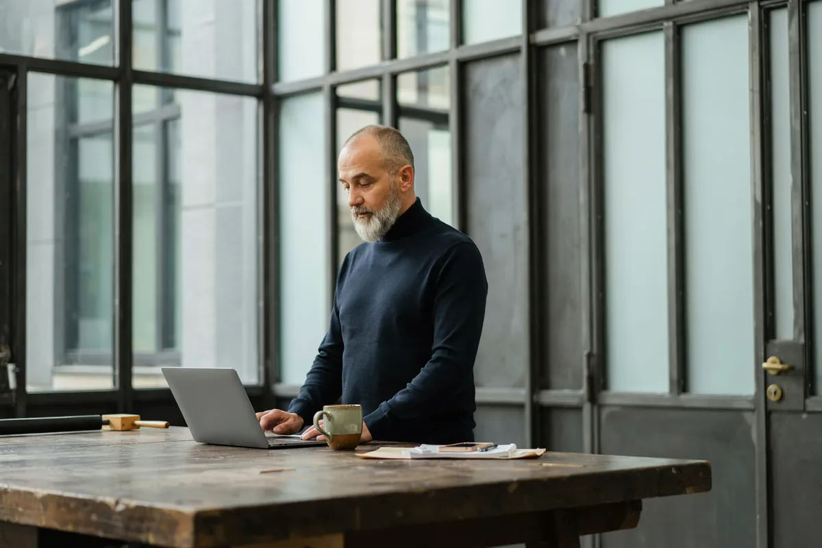 Neuausrichtung im digitalen Wandel: Videocoaching Mann mit grauweißem Bart im Rollkragenpullover steht im hellen Atelier am Holztisch und arbeitet konzentriert am Laptop