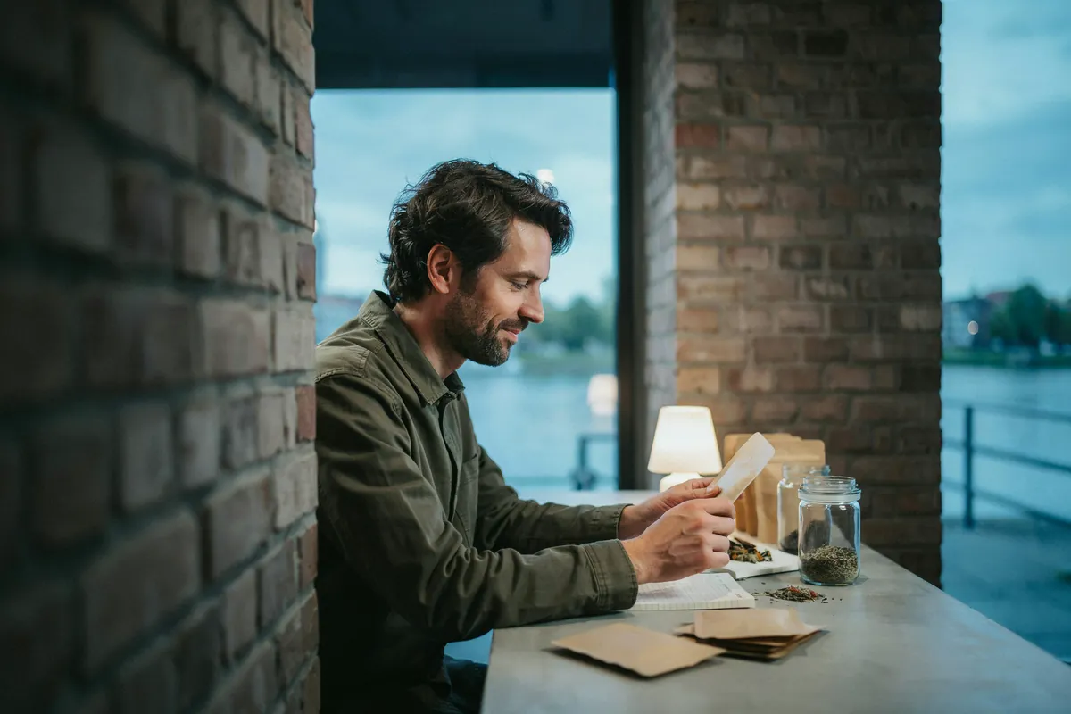 E-Commerce Coaching: Gründung im Onlinehandel Mann mit Bart sitzt im Backsteinloft am Fenster zum Fluss, hält eine Teetüte und lächelt, daneben Gläser mit losem Tee.