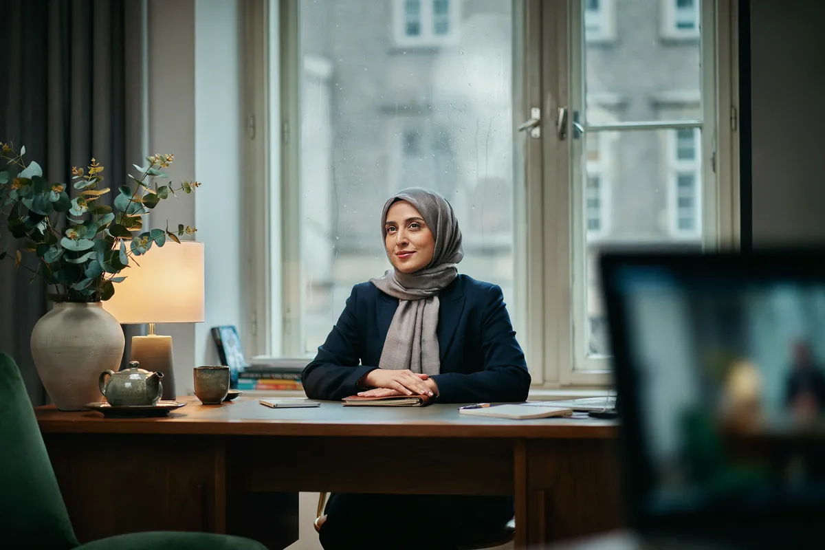 Frau mit grauem Hijab sitzt im Videocoaching am Nussbaumschreibtisch und lächelt ruhig, am Fenster laufen Regentropfen herab.
