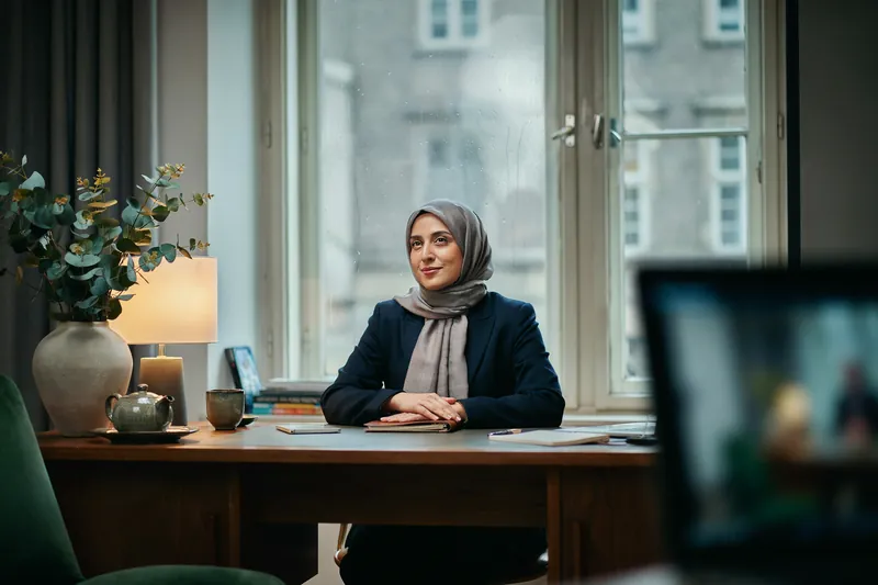 Frau mit grauem Hijab sitzt im Videocoaching am Nussbaumschreibtisch und lächelt ruhig, am Fenster laufen Regentropfen herab.