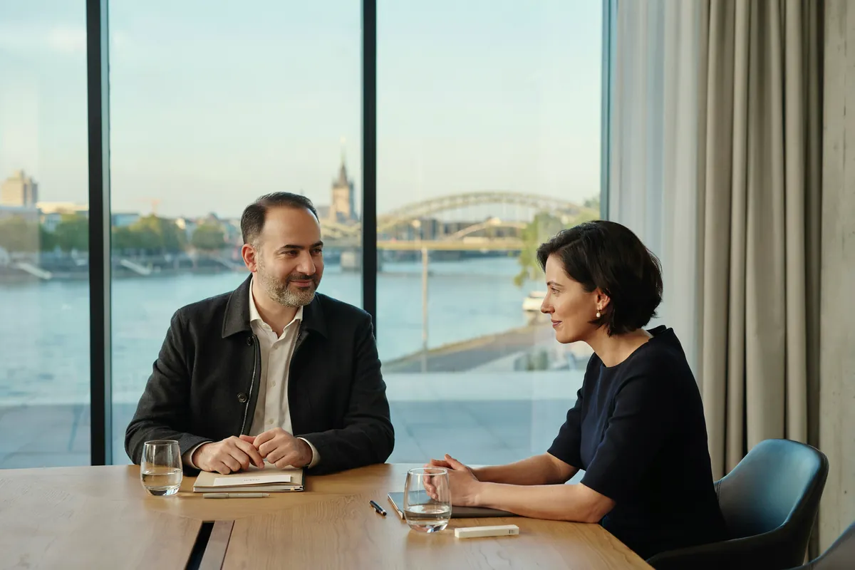 Integrationscoaching: Gespräch über dem Rhein Mann mit grauem Bart sitzt im Glasraum über dem Rhein am Tisch, seine Coachin hört ihm lächelnd zu.
