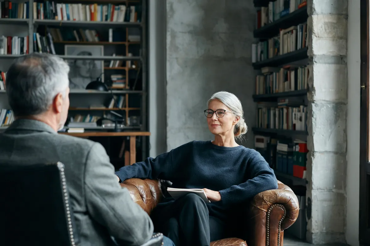 Frau mit silbernem Haarknoten und Brille sitzt lächelnd im Ledersessel vor Bücherregalen, ihr Coach sitzt ihr gegenüber