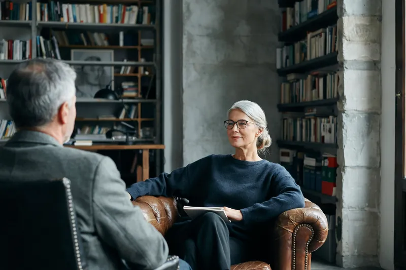 Frau mit silbernem Haarknoten und Brille sitzt lächelnd im Ledersessel vor Bücherregalen, ihr Coach sitzt ihr gegenüber