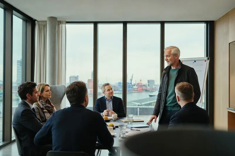 Mann mit grauem Haar steht im Besprechungsraum mit Hafenblick vor einem Flipchart, eine Kollegin und vier Kollegen hören zu.