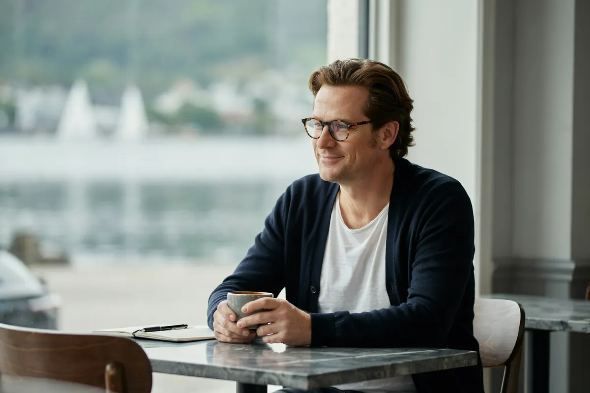 Online Marketing Advanced: der fundierte Wechsel Mann mit runder Brille sitzt mit Kaffeetasse und Notizbuch am Marmortisch im Café und lächelt, draußen die Kieler Förde.