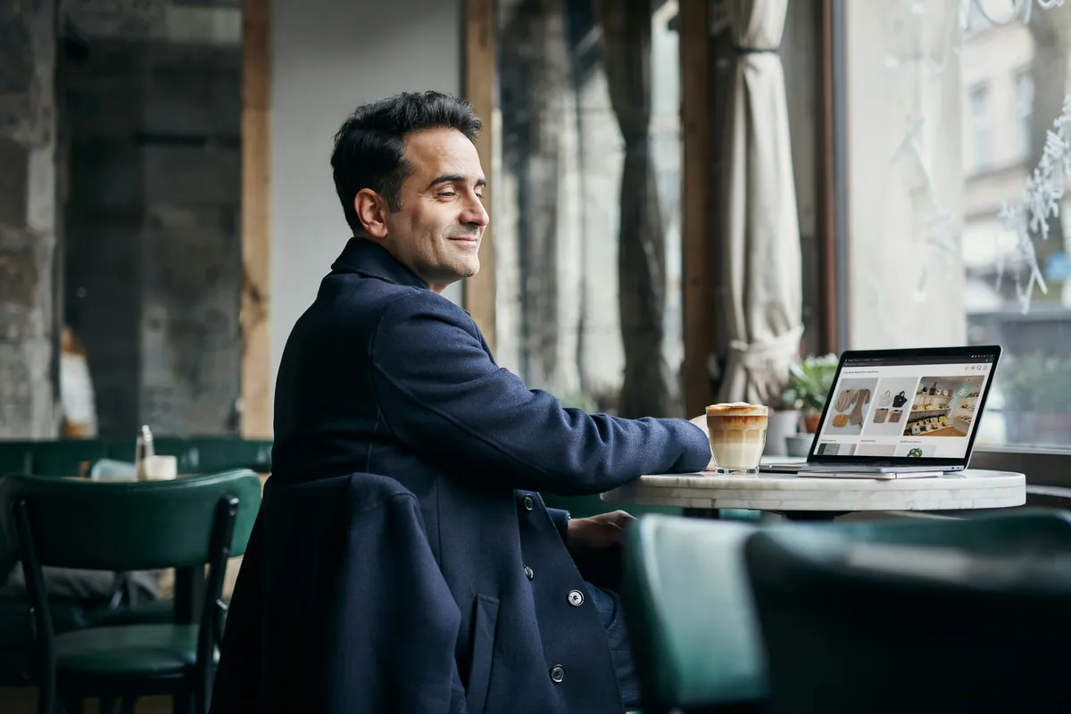 Dunkelhaariger Mann im Wollmantel sitzt an einem Marmortisch in einem Café in Haidhausen vor seinem Laptop und lächelt.