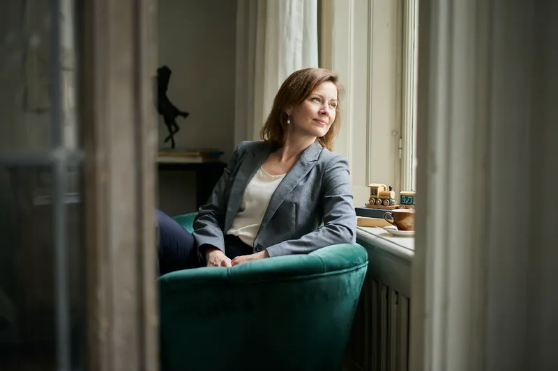 Frau mit kastanienbraunem Haar sitzt im grünen Samtsessel am Fenster einer Altbauwohnung und blickt lächelnd hinaus