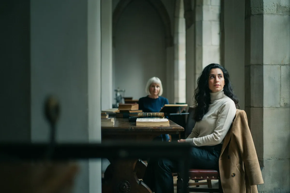 Frau mit langem dunklem Haar sitzt nachdenklich in einer historischen Bibliothek mit Steinbögen, hinten am Tisch ihre Coachin