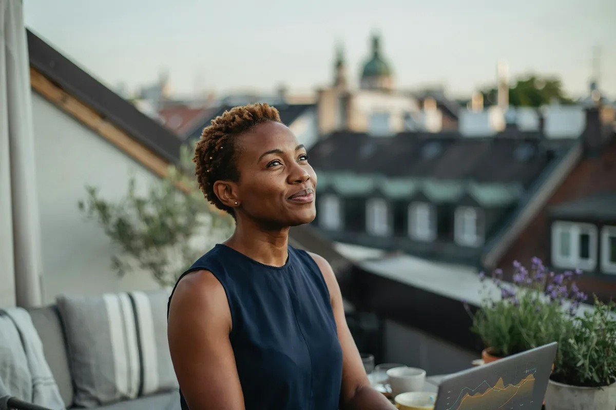 Frau mit kurzem Naturhaar blickt auf einer Dachterrasse in Schwabing neben ihrem Laptop lächelnd in die Ferne.