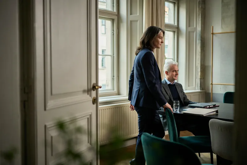 Unternehmerin im dunkelblauen Anzug steht am Fenster eines hellen Altbau-Sitzungsraums, am Tisch sitzt lächelnd ihr Coach.
