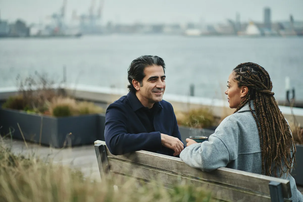 Stressmanagement und Burn-out-Prophylaxe im Coaching Mann mit dunklem, an den Seiten ergrautem Haar sitzt entspannt neben seiner Coachin auf einer Dachterrasse über dem Hafen.