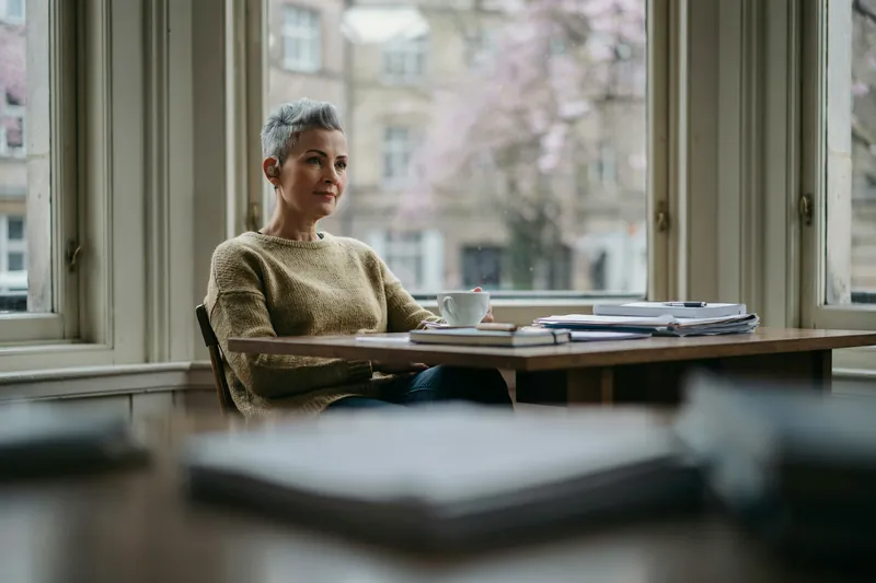 Frau mit kurzem grauem Haar und Hörgerät sitzt mit Teetasse am Schreibtisch vor ihren Unterlagen, draußen blühen Kirschbäume.