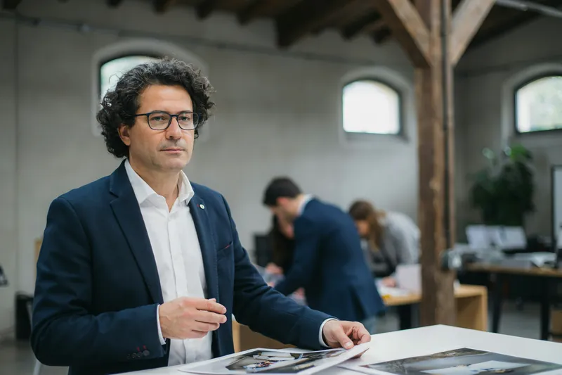 Mann mit dunklen Locken und Brille steht in einer Halle mit Holzbalken am Tisch mit Ausdrucken, dahinter ein Team.