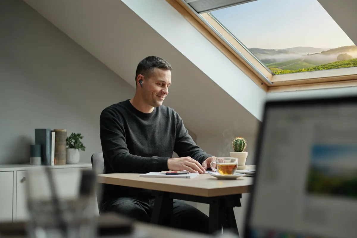 Mann mit kurzem dunklem Haar und Ohrhörern folgt im Dachzimmer einem Live-Onlinekurs am Laptop, draußen Weinberge im Dunst.