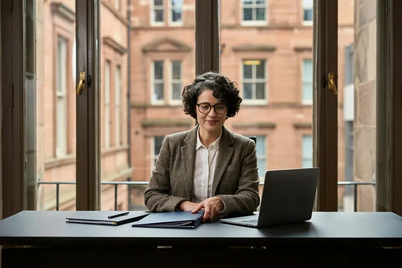 Frau mit kurzen dunklen Locken und Brille legt am Schreibtisch die Hand auf eine Mappe, dahinter rote Sandsteinfassaden.